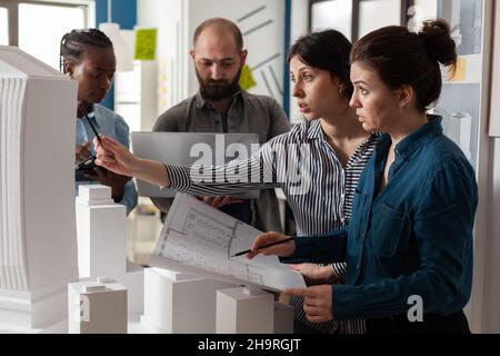 Two women professional architects holding blueprints next to colleagues holding laptop looking at construction plans. Group of focused architectural engineers analyzing white foam scale model. Stock Photo