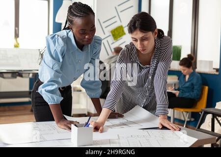 Focused team of women architects inspecting construction plans standing at desk in modern architectural office. Team of two engineers colleagues discussing blueprints for residential project. Stock Photo