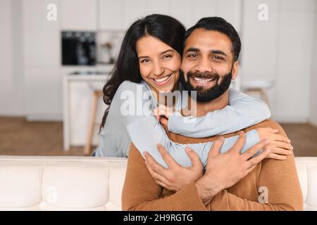 Happy lovely young Indian couple together at home, ethnic young wife hugging from behind her husband, sitting and resting on sofa in modern apartment, portrait of romantic multiracial couple in love Stock Photo