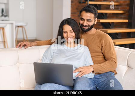 Serene Indian couple spending time together at home, using laptop for online shopping, web browsing, ethnic young wife and husband resting on the sofa in modern apartment, watching movie together Stock Photo