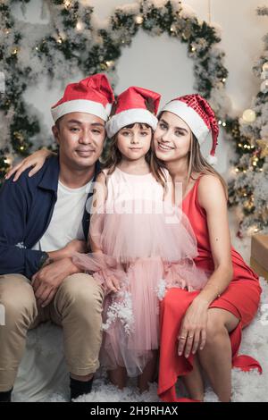 Christmas studio family shoot in red Christmas hats. New Year photo of happy mixed family with cute daughter. Everyone smile Stock Photo