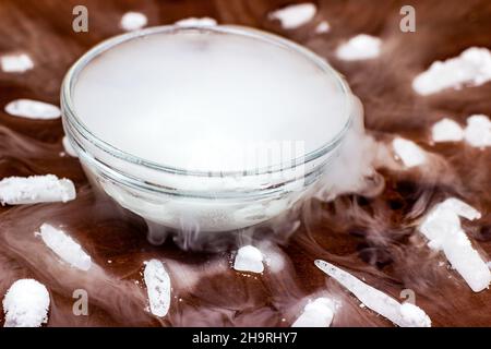 White cool dry ice (frozen carbon dioxide) with smoke effect in the glass bowl on the dark brown wooden background. Stock Photo