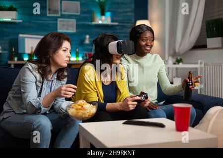 Concentrated woman with virtual reality headset holding controller playing videogames while multi-ethnic friends helping her during online competition enjoying spending time together. Hanging out concept Stock Photo
