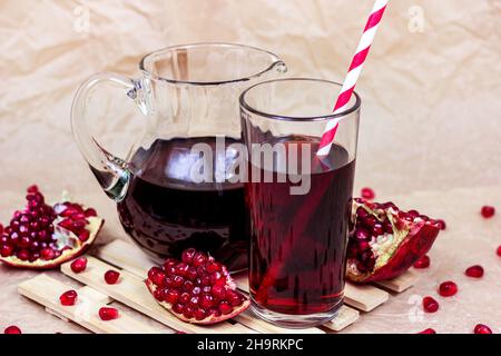 Red pomegranate juice in a glass with a straw and jar with garnet pieces on light wooden background. Stock Photo