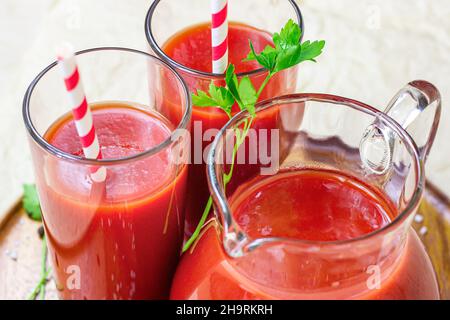 Fresh red tomato juice in a glass with a straw and jar with tomatoes on light wooden background. Stock Photo