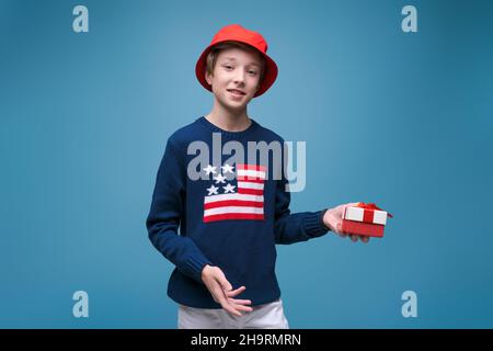 Joyful boy with holiday surprise gift celebrating holiday on blue studio background at birthday party. Happy laughing guy with a real box in his hands, in a blue sweater with a usa flag Stock Photo