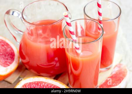 Fresh red grapefruit juice in a glass with a straw and jar with fruit pieces on light wooden background. Stock Photo