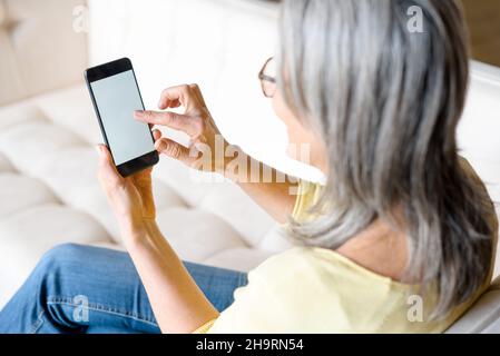 Back view a woman holding smartphone with an empty screen, senior gray-haired woman sits on couch and holds gadget with blank display, touching touchscreen with finger, mock-up, app advertising Stock Photo