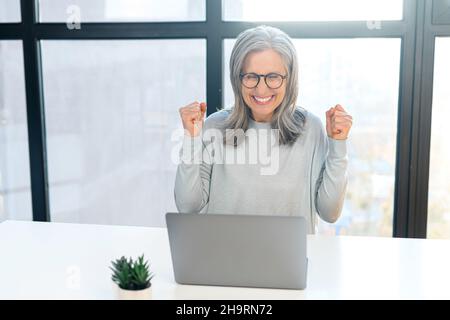Overjoyed mature woman in casual wear with laptop in office, watching at the screen and screaming happily, excited senior lady got job hiring, get good news or won in lottery. Victory concept Stock Photo