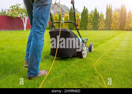 A gardener with a lawn mower is cutting green grass in the garden, in the house backyard on a sunny summer day. The legs of the worker and a lawn mowe Stock Photo