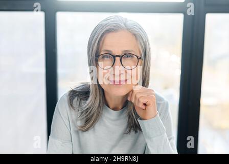 Positive senior businesswoman wearing stylish glasses looks at camera and smiles. Close-up portrait of confident gray-haired woman, mature female teacher, ceo, experienced manager Stock Photo