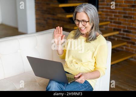 Smiling mature woman wearing a yellow jumper is sitting in the living room, relaxing and talking to her friend or family, waving at the laptop screen, staying connected during pandemic Stock Photo