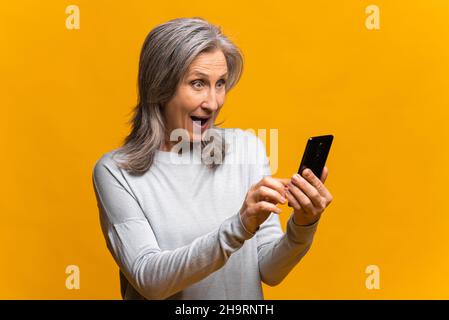 Astonished senior woman holding and using smartphone, surprised mature modern gray-haired lady staring at phone screen, receiving unexpected good news, crazy sale deal, isolated over yellow background Stock Photo