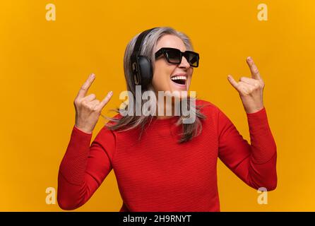 Happy crazy elderly mature woman in sunglasses and headphones making horn signs isolated on yellow, overjoyed senior gray-haired female in red jumper showing rock symbol, laughing and singing Stock Photo