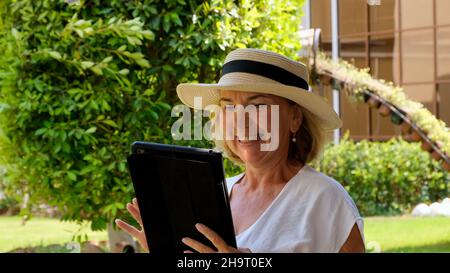 Senior female blonde businesswoman in hat uses tablet pc, digital tablet for business work or study in her own green garden. woman aged 50-55. The Stock Photo