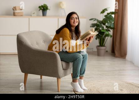 Happy young calm muslim woman sitting on comfy armchair and reading paper book, enjoying morning and smiling Stock Photo
