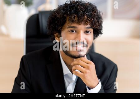 Close-up business portrait. Friendly smiling curly-haired Indian business man, small business owner, company leader or sales manager, in formal suit, successful real estate agent, looking at camera Stock Photo