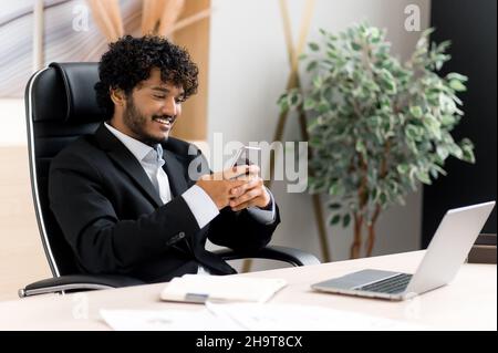 Remote communication using a smartphone. Indian positive man sitting at his desk in the office, texting online his gadget, answers email, browses the internet, smiling friendly Stock Photo