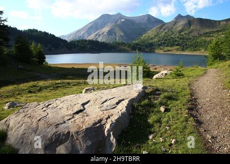 A footpath runs along a large flat rock in front of the Lac d'Allos with Notre Dame des Monts on the far side of the lake (Parc du Mercantour) Stock Photo