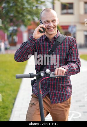 Young man in casual trousers and shirt standing next to his electric scooter, talking on the mobile phone, smiling Stock Photo