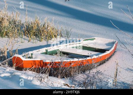 Wooden colorful boat stacked on a frozen lake covered by snow on the beach with reeds Stock Photo