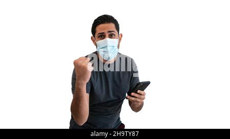 Caucasian man using smartphone with sterile face mask isolated on white background studio. Guy celebrating success at quarantine pandemic coronavirus Stock Photo