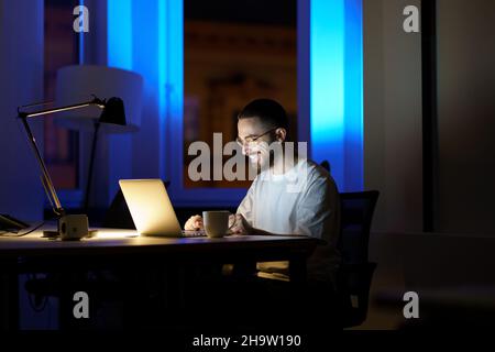 Smiling man talk using online video call in dark office stay overtime to communicate with coworkers Stock Photo