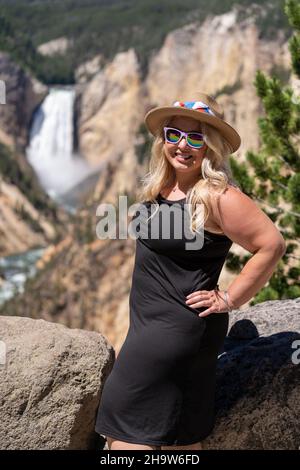 Beautiful cute blonde woman poses with her hands on hips at the waterfall at the Grand Canyon of Yellowstone National Park Stock Photo