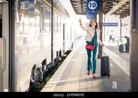 Beautiful girl running and chasing the leaving train in station. Waving hand and rushing to get on - Young business woman with suitcase running Stock Photo