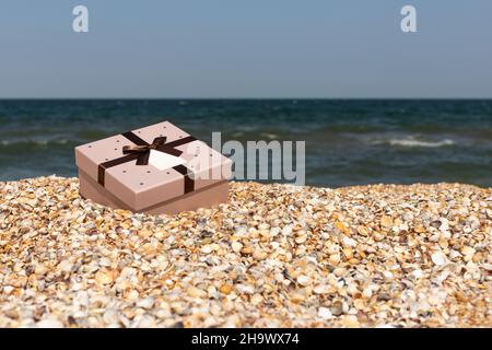 A box with a gift tied with a ribbon lies on the beach near the sea. Front view, vertical. Stock Photo