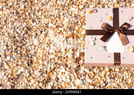A box with a gift tied with a ribbon lies on the beach near the sea. Front view, vertical. Stock Photo