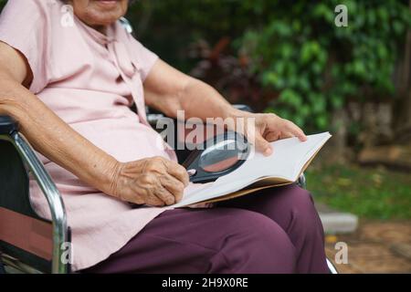 Elderly woman using a magnifying glass to read on wheelchair. Stock Photo