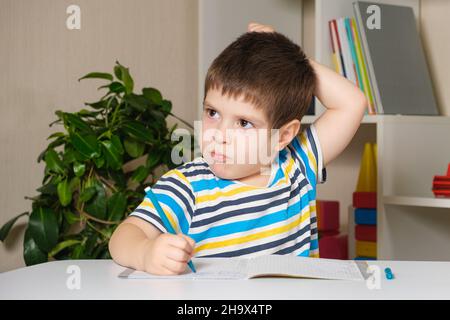 A boy in a striped T-shirt scratches his head, writes spells, prepares for school. Stock Photo