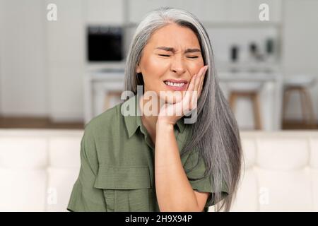 Troubled mature 50s woman with gray-hair feels strong toothache, holding cheek and suffering from strong dental pain sitting on sofa, middle-aged female undergoing tooth pain, enamel sensitivity Stock Photo