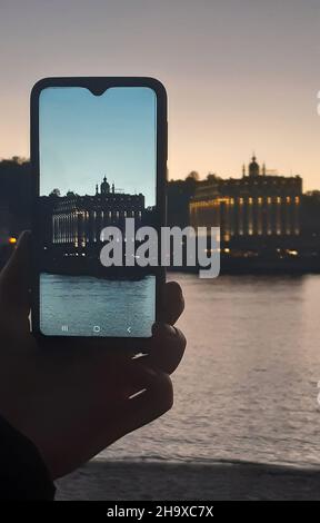 A human hand photographs a building across the river on a mobile phone. The building is clear on the phone,and the building is blurry in the backgroud Stock Photo