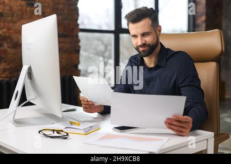Smiling male office employee doing paperwork sitting at the desk, businessman checking annual report, manager reading contract sitting in front of PC, smart bearded guy in shirt doing routine work Stock Photo