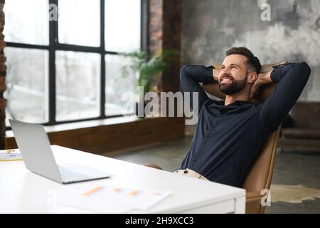 Take a break on a workplace. Young male office employee leaned back in chair relaxed, pleased with the work done guy resting sitting at the desk in loft office, satisfied guy daydreaming Stock Photo