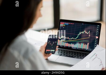 Female crypto trader, investor, using laptop and cellphone analyzing digital cryptocurrency exchange stock market trading graphs, doing analysis, thinks about the future of the crypto market Stock Photo