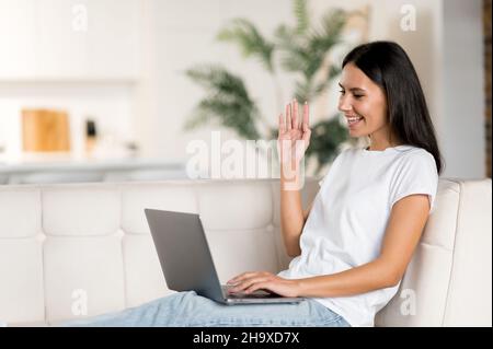 Happy caucasian young woman uses laptop, having a video call, communicate with colleagues or friends by video conference, waving hand and smiling. Pretty brunette woman sits on a couch in living room Stock Photo