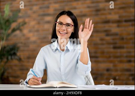 Close-up webcam headshot, a friendly pretty smart young adult caucasian woman, top manager, financial advisor, sits at work desk, talking with colleagues, partners via video link, waving hand, smiles Stock Photo