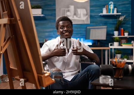 Portrait of smiling young artist in wheelchair holding graphic pencil drawing sketch on canvas working at artistic masterpiece in art studio. Illustrator with disability painting. Creativity concept Stock Photo