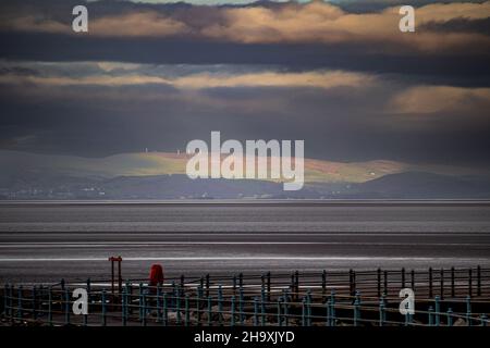 Morecambe, Lancashire, United Kingdom. 9th Dec, 2021. Sunshine breaks through the clouds over Morcambe Bay to illuminate Kirkby Moor in the South Lakeland Fells Credit: PN News/Alamy Live News Stock Photo