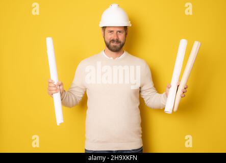 Young smiling architect man wearing contractor helmet holding project paper plan over isolated yellow background Stock Photo