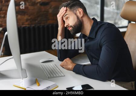 Depressed young male office employee is staring at computer monitor with despair, worried businessman made mistake in project, fail deadline Stock Photo