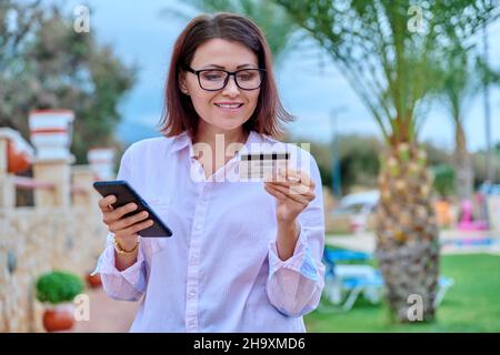 Middle age smiling woman with credit card and smart phone in hands, outdoor Stock Photo