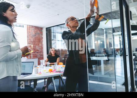 Creative businesswomen sticking adhesive notes to a glass wall with their colleagues in the background. Two focused businesswomen sharing their ideas Stock Photo