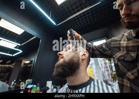Barber works with a beard clipper. Hipster client getting haircut. Stock Photo