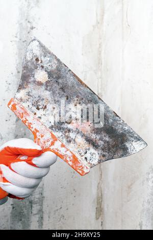 Plasterer holds an old special scraper in his hand on background of gray concrete wall. Home redecoration during pandemic. Close-up view, selective Stock Photo