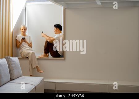 Couple using smartphones on windowsill at home Stock Photo