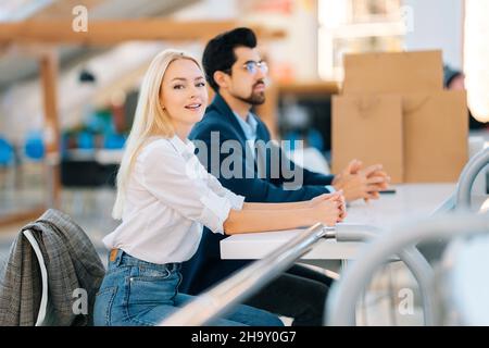 Happy beautiful young man and woman waiting coffee, resting sitting at table in mall after shopping at mall. Stock Photo
